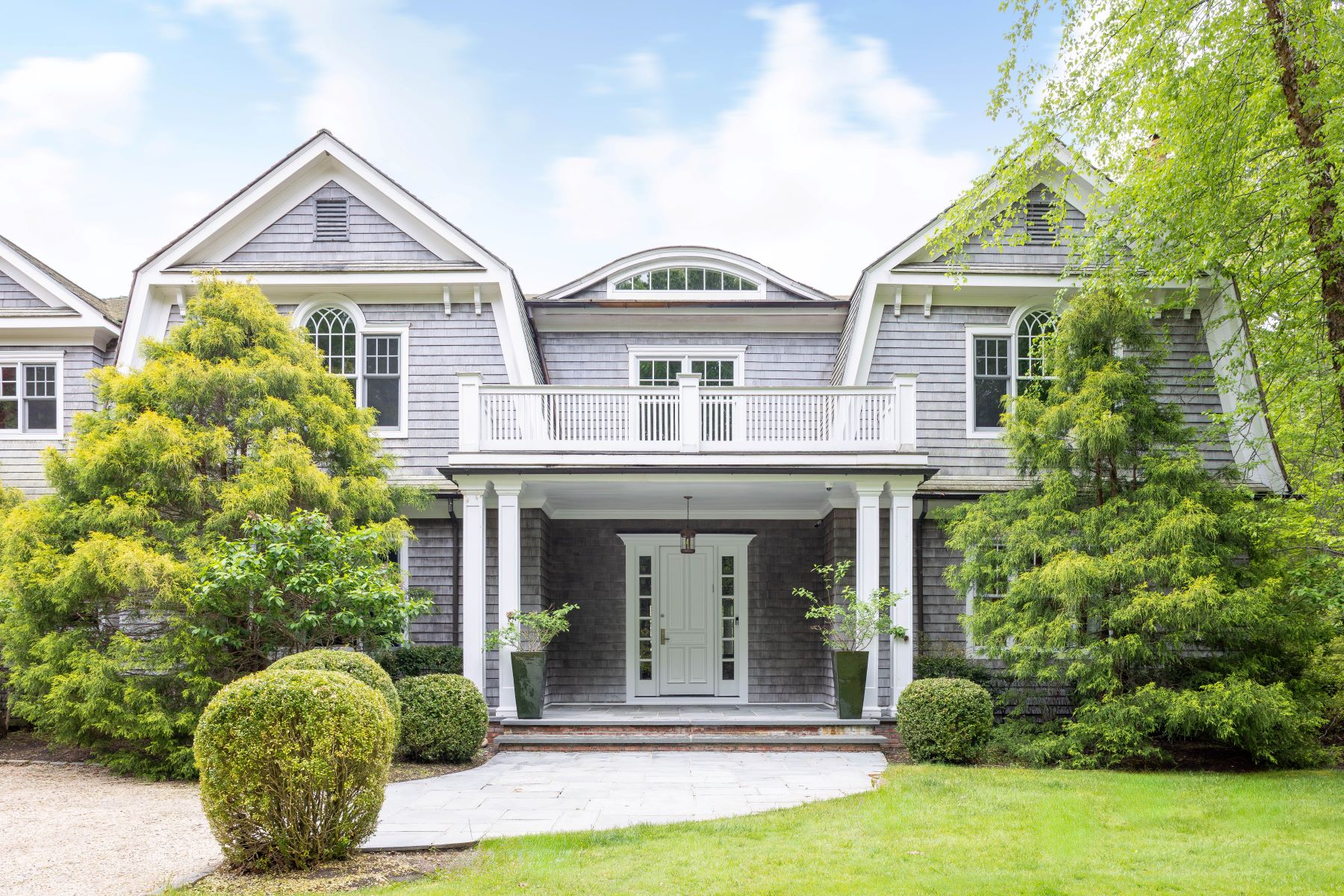 The front entrance of the estate: a gray cedar-shingle facade with a gambrel roofline, a second-story balcony over a columned covered porch, flanked by sculpted evergreens and boxwood