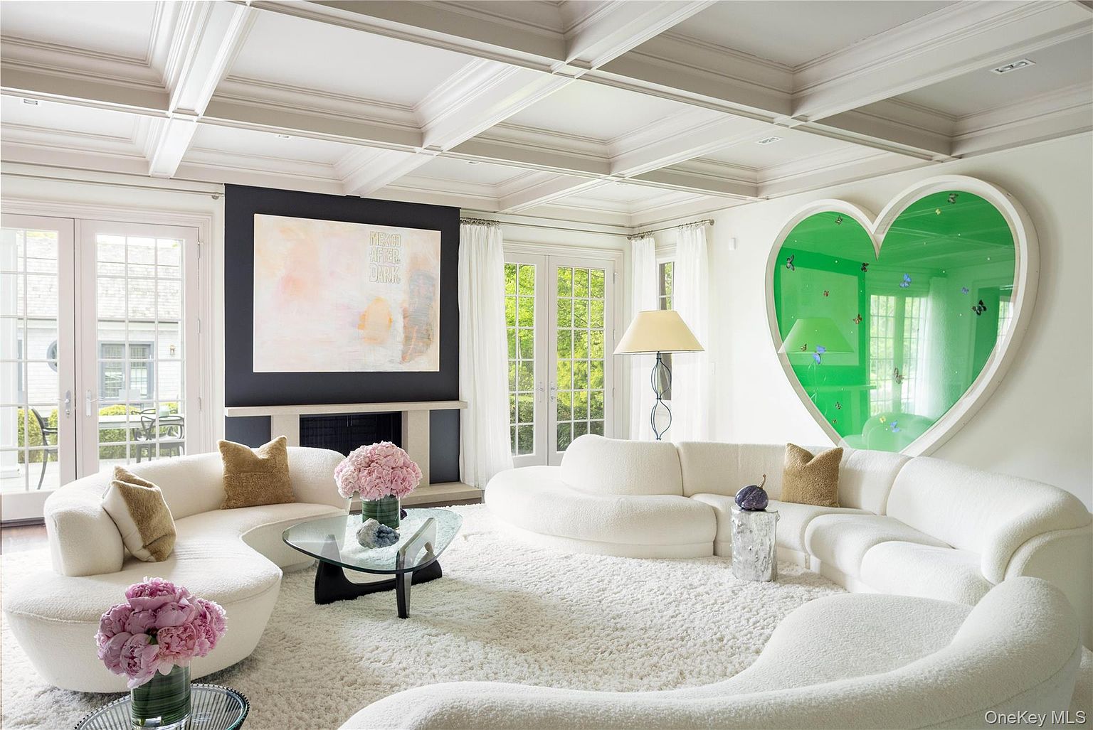 A coffered-ceiling living room with curving white boucle sofas around a glass cocktail table, a fireplace flanked by French doors, and a backlit emerald-green heart-shaped art niche set into the wall