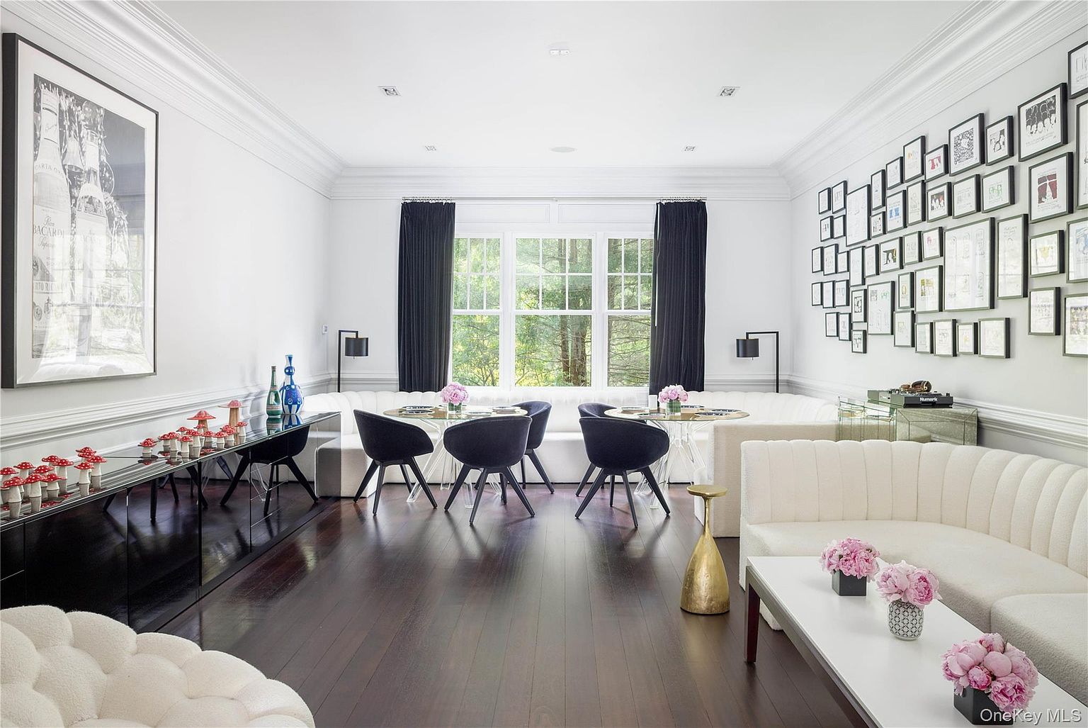 A dining and sitting room with dark hardwood floors, a salon-style wall hung with dozens of framed works, black velvet dining chairs around a glass table, white tufted banquettes and pink peonies