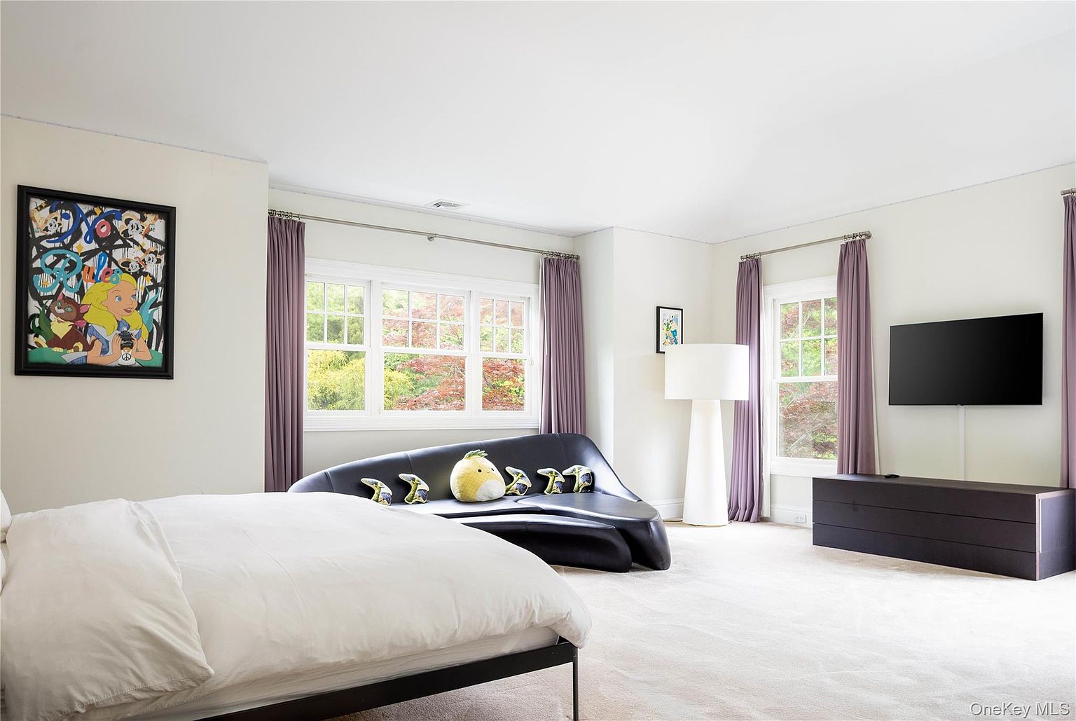 A bright primary bedroom with pale walls, a low platform bed, a sculptural black leather chaise, a pop-art canvas, lavender drapes framing two windows, and soft carpet underfoot