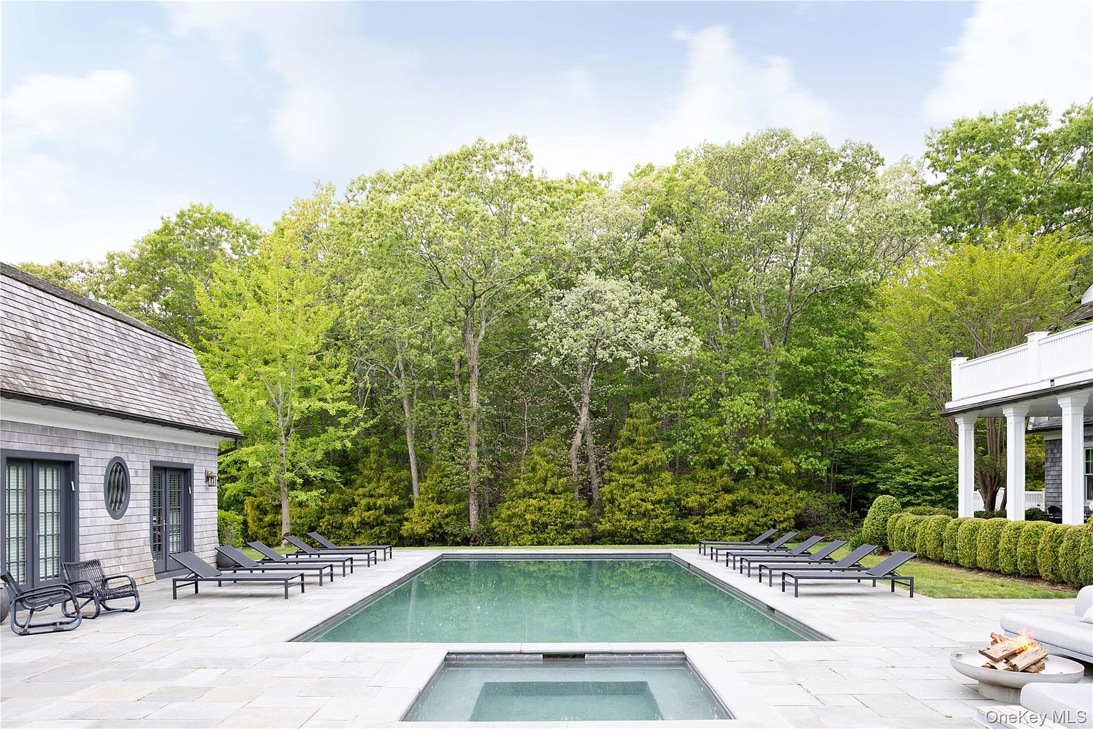 A long rectangular pool with a sunken spa at one end, bluestone surround lined with chaise lounges, a sunken fire feature, and a shingled pool house set against a wall of mature trees