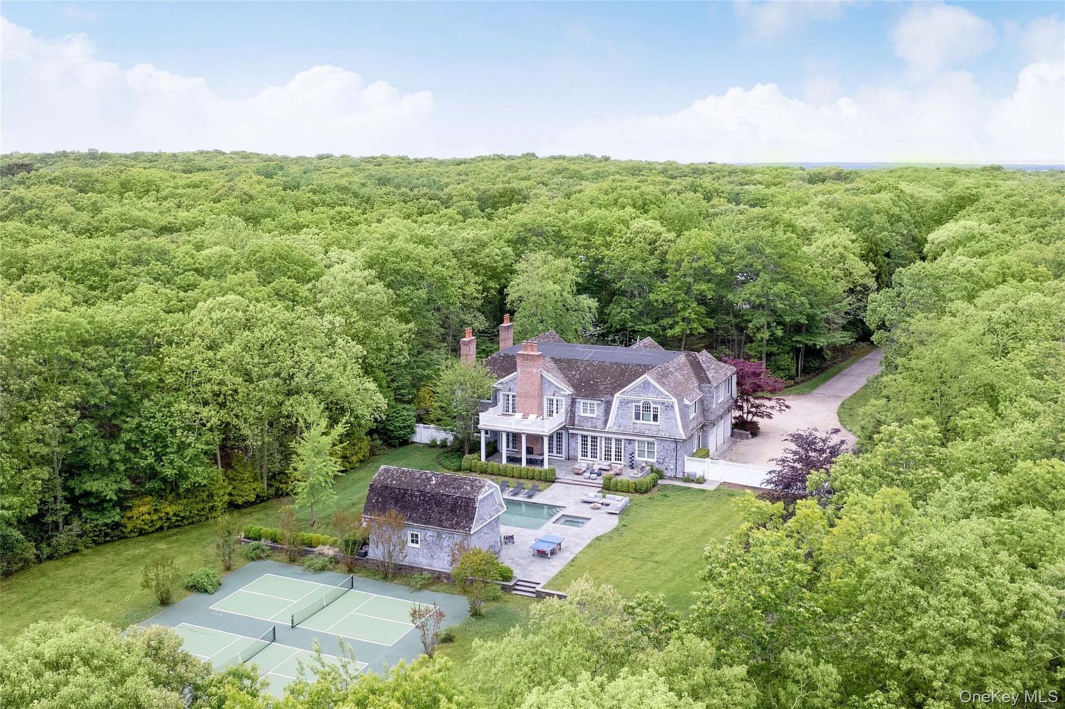 An aerial three-quarter view of the estate showing the main house and pool beside a separate barn-form building, a tennis-lined sport court and pickleball courts, all set in a clearing surrounded by dense woodland