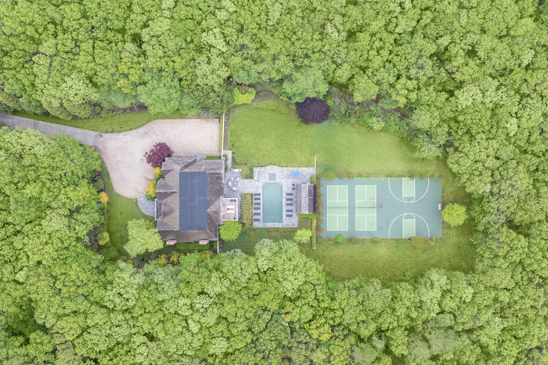 A directly overhead aerial of the full parcel: the main residence and outbuildings, a green sport court and a blue-green pickleball court, gravel drive and expansive lawns enclosed entirely by mature trees