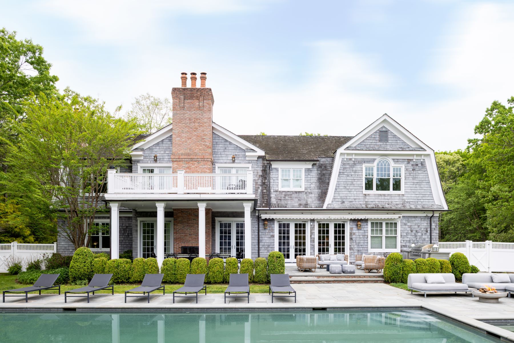 The rear elevation of 486 Middle Line Highway, a gray shingle-style Hamptons estate with a brick chimney and covered loggia, seen across the bluestone pool terrace lined with lounge chairs